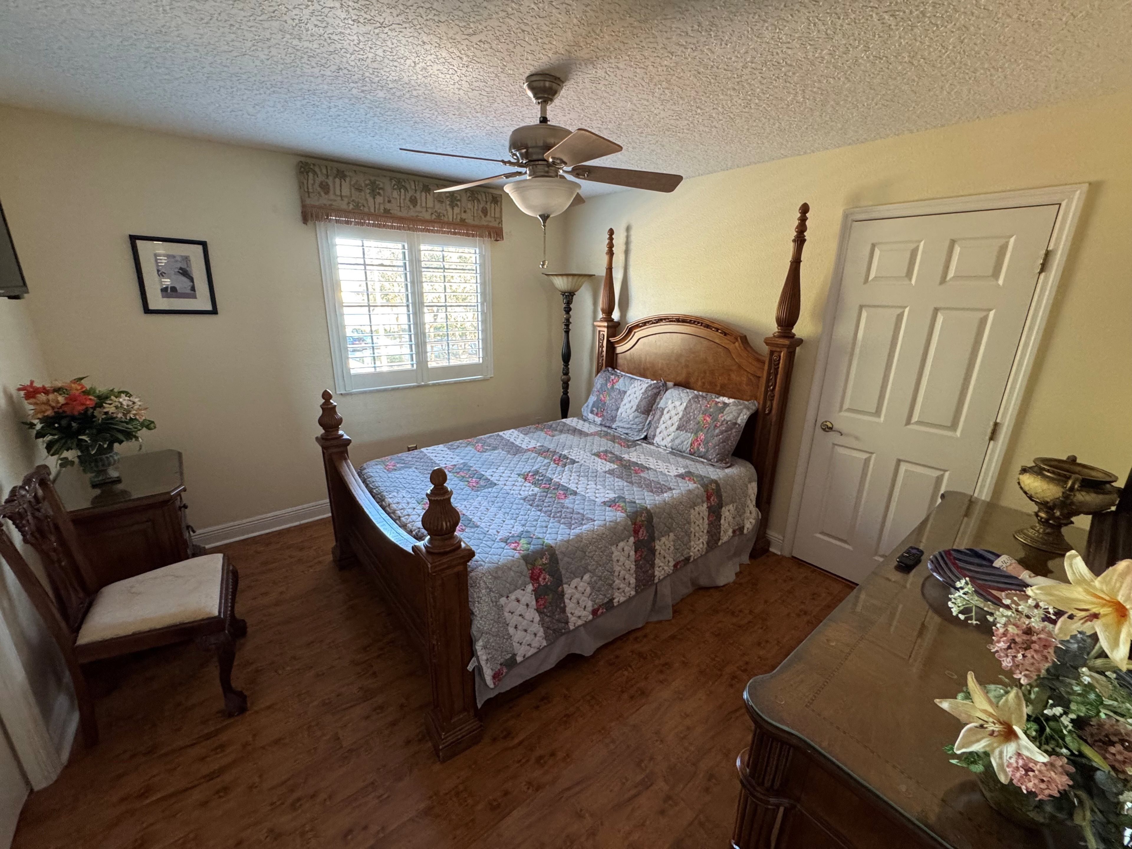 A bedroom with a wooden four-poster bed, a ceiling fan, a window with blinds, a door, and a small table with a vase of flowers.