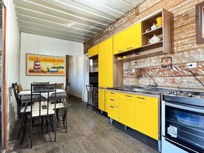 A kitchen with yellow cabinets, a wooden backsplash, and a painting of a lighthouse. - Contêiner Exclusivo - Praia da Gamboa (Garopaba)