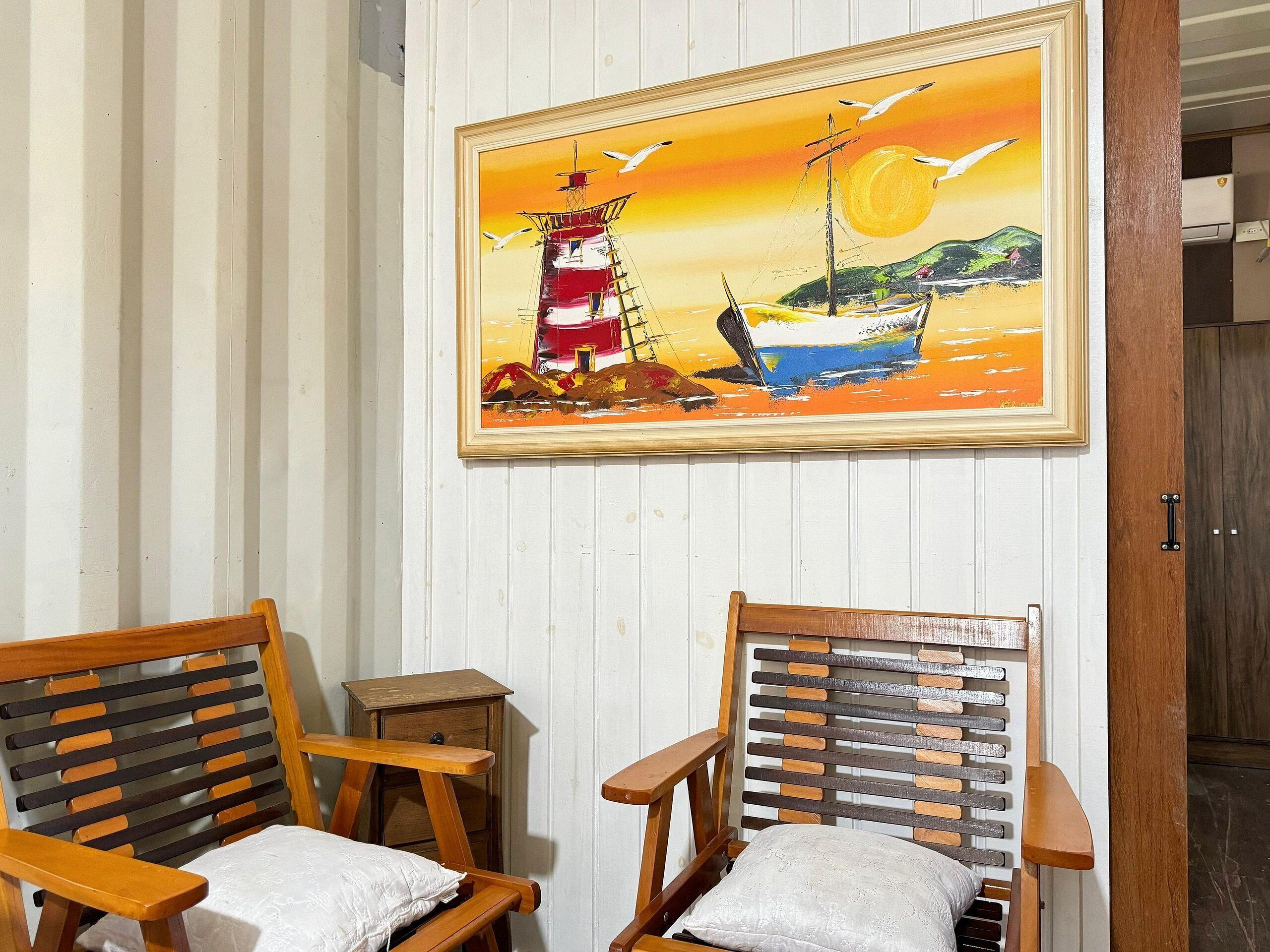 A room with two wooden chairs, a small wooden table, and a framed painting of a lighthouse and sailboat on the wall.