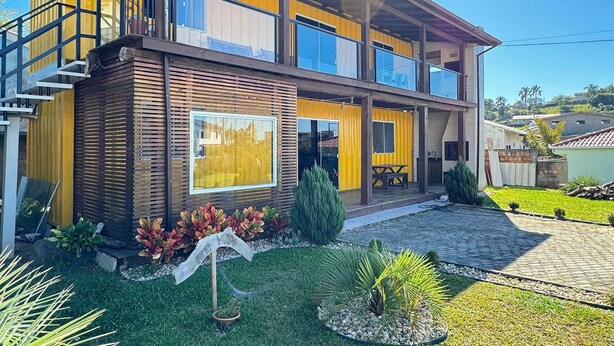 A two-story house with a balcony, a wooden exterior, and a garden with plants and a sign. - Contêiner Exclusivo - Praia da Gamboa (Garopaba)