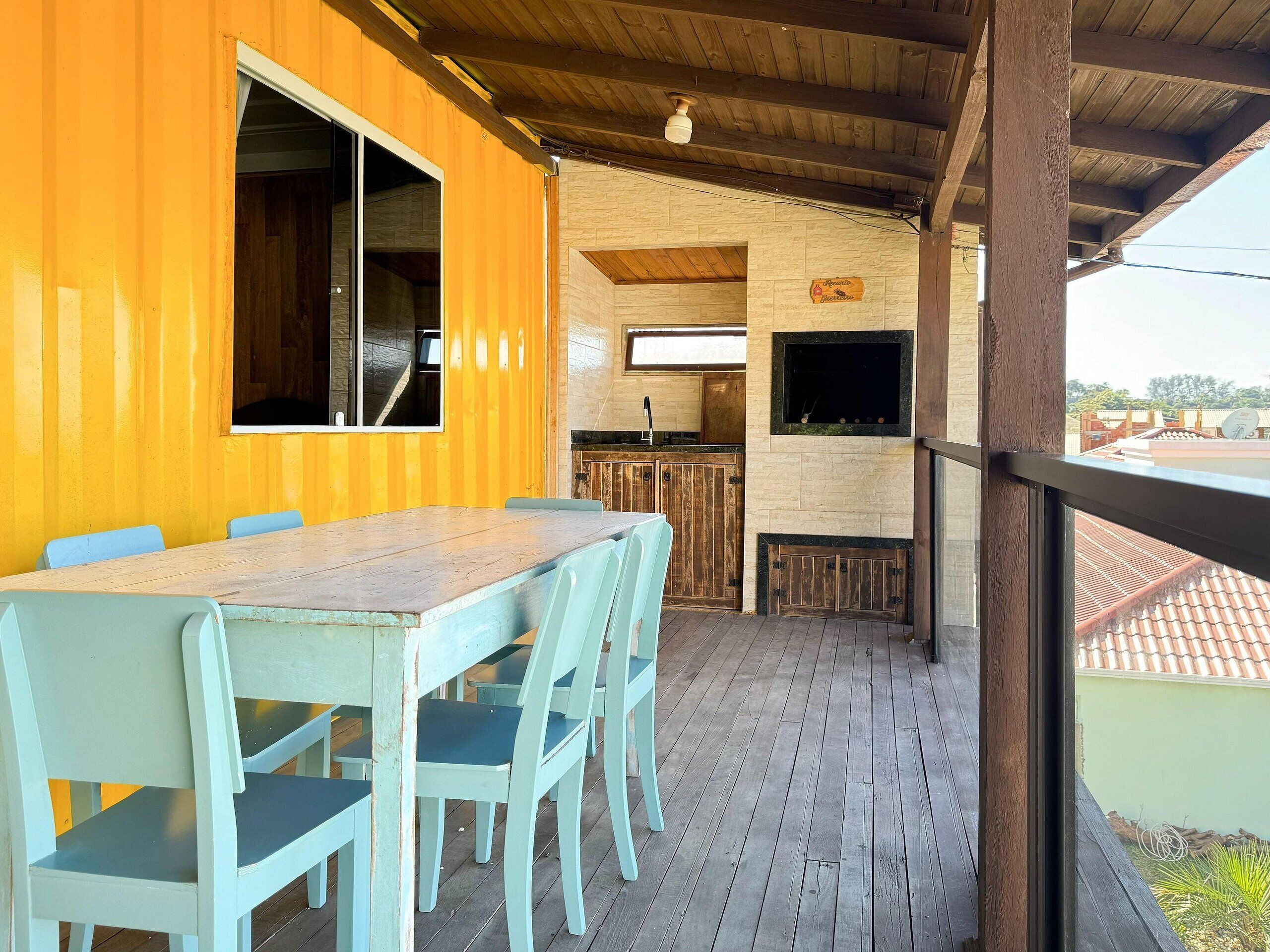 Um deck de madeira com mesa e cadeiras, uma área de cozinha com parede de pedra e vista para os arredores.