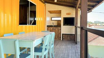 Um deck de madeira com mesa e cadeiras, uma área de cozinha com parede de pedra e vista para os arredores.