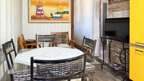 A dining area with a round table, chairs, and a painting of a sunset over a lighthouse. - Contêiner Exclusivo - Praia da Gamboa (Garopaba)