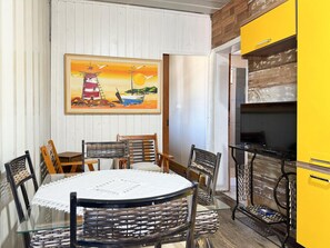 A dining area with a round table, chairs, and a painting of a sunset over a lighthouse. - Contêiner Exclusivo - Praia da Gamboa (Garopaba)
