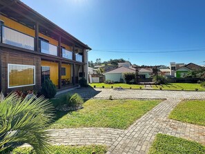 A modern two-story building with large windows, a covered patio, and a paved courtyard with grass and pathways. - Contêiner Exclusivo - Praia da Gamboa (Garopaba)