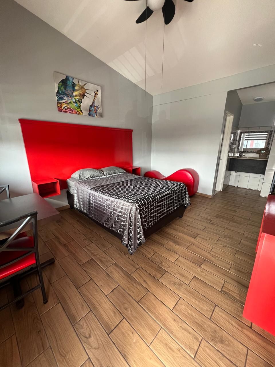 Un dormitorio moderno con cabecera roja, cama con colcha estampada, suelo de madera y una zona de comedor con mesa y sillas rojas.