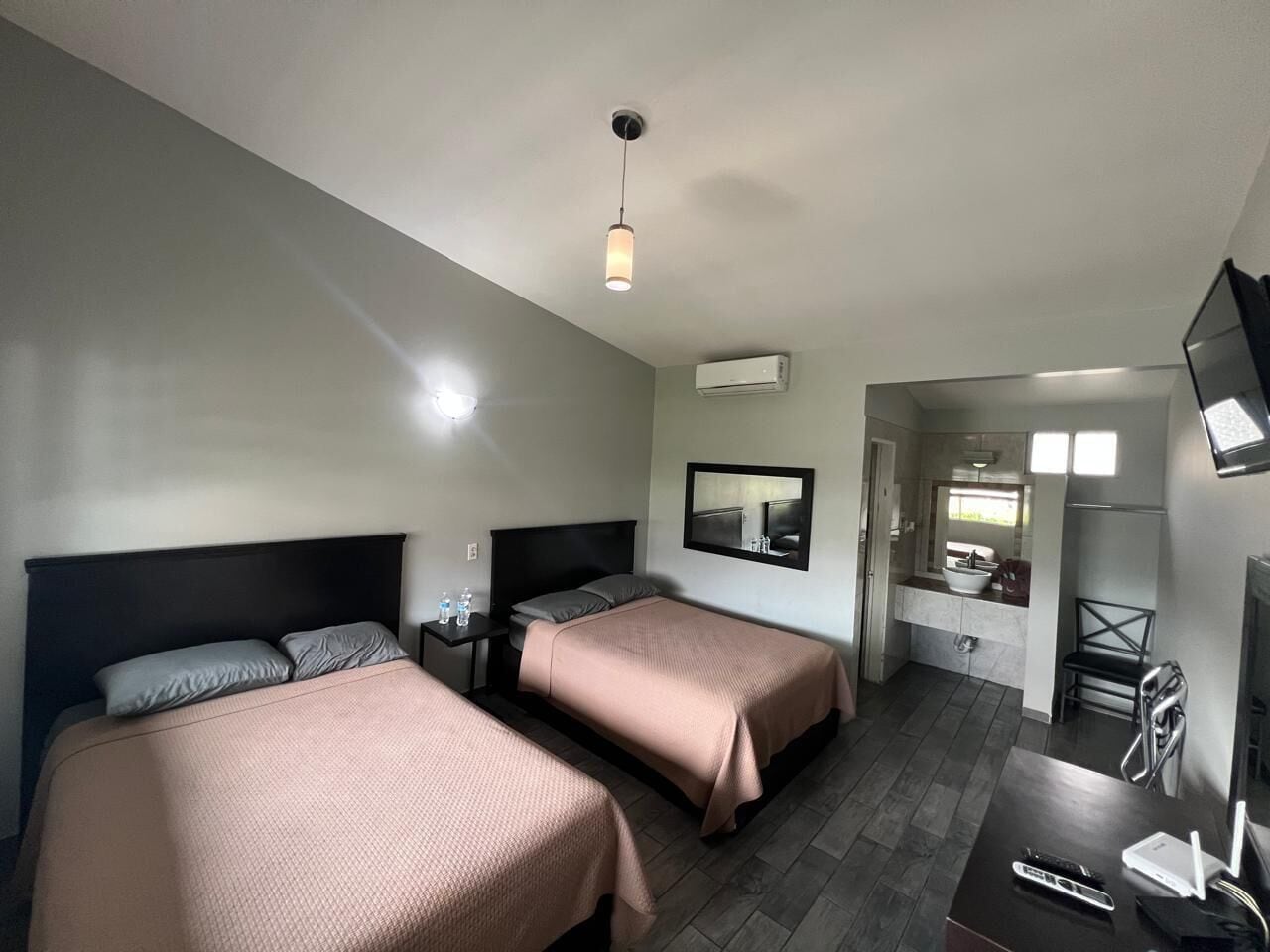 A hotel room with two beds, a desk, a chair, a TV, and a bathroom with a sink and mirror.