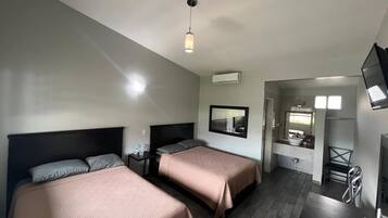 A hotel room with two beds, a desk, a chair, a TV, and a bathroom with a sink and mirror.