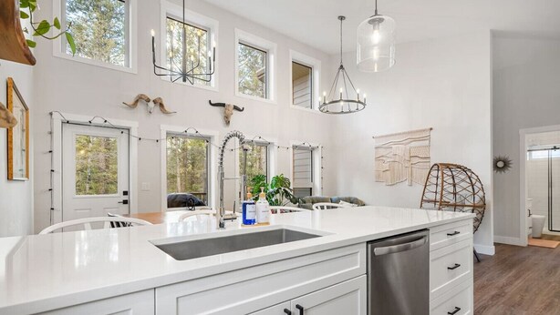Una cocina moderna con armarios blancos, un lavavajillas de acero inoxidable y una isla grande con un lavamanos. Varias ventanas proporcionan luz natural y hay decoración en la pared sobre la isla.