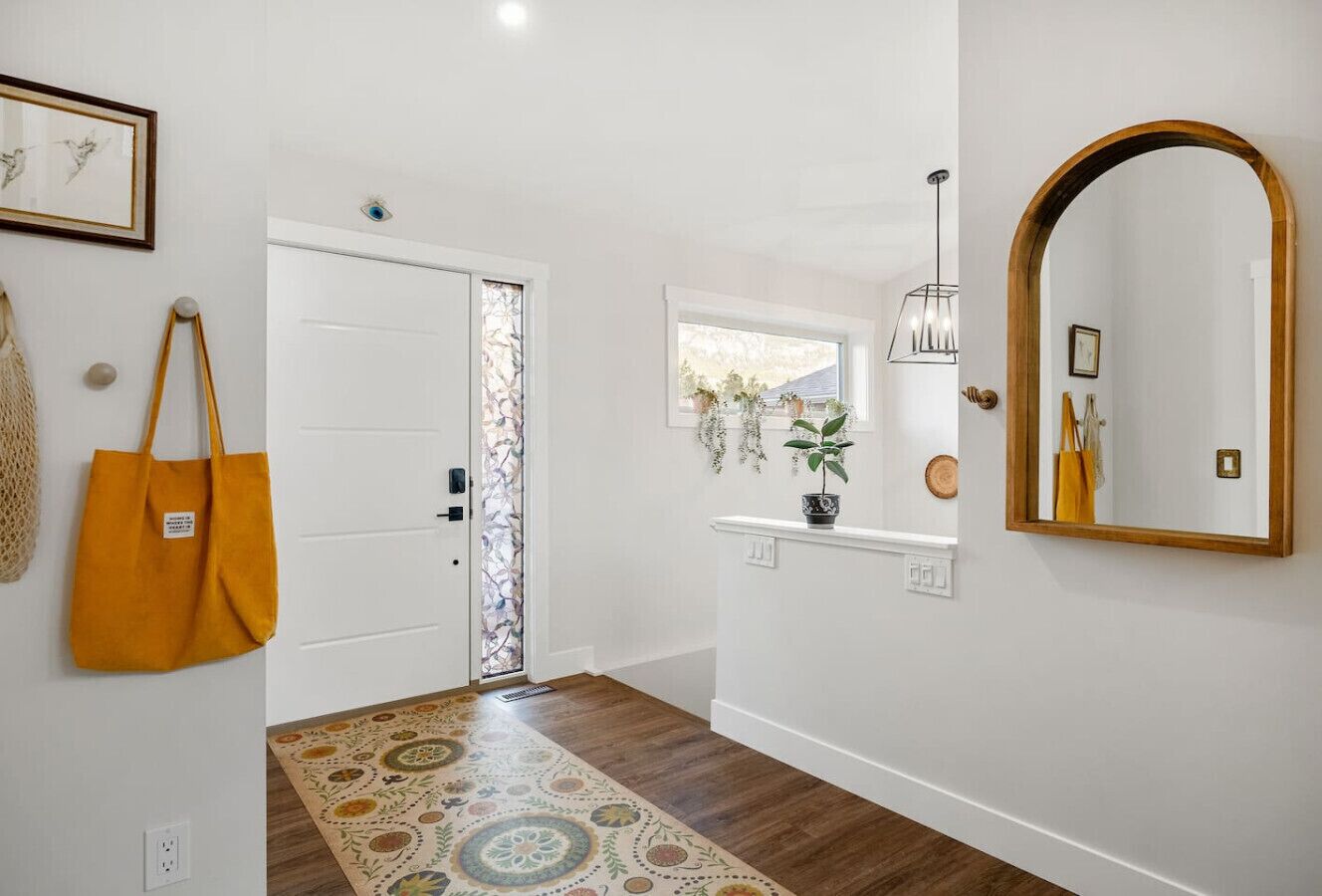 A hallway with a patterned rug, a white door, a small window with plants, and a mirror on the wall.