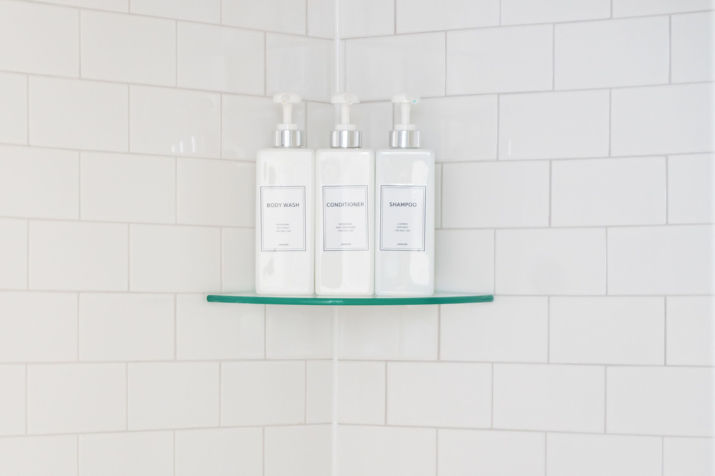 Three white bottles labeled BODY WASH, CONDITIONER, and SHAMPOO on a glass shelf against a white tiled wall.