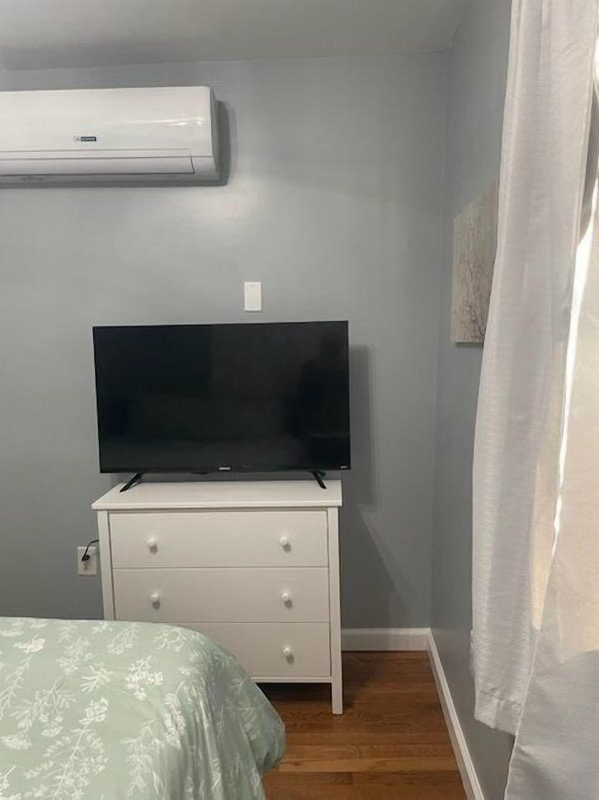 Camera da letto con TV a schermo piatto su una cassettiera bianca, un letto con copripiumino verde con motivo floreale e un condizionatore d'aria a parete.