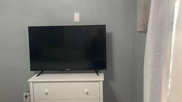 Camera da letto con TV a schermo piatto su una cassettiera bianca, un letto con copripiumino verde con motivo floreale e un condizionatore d'aria a parete.