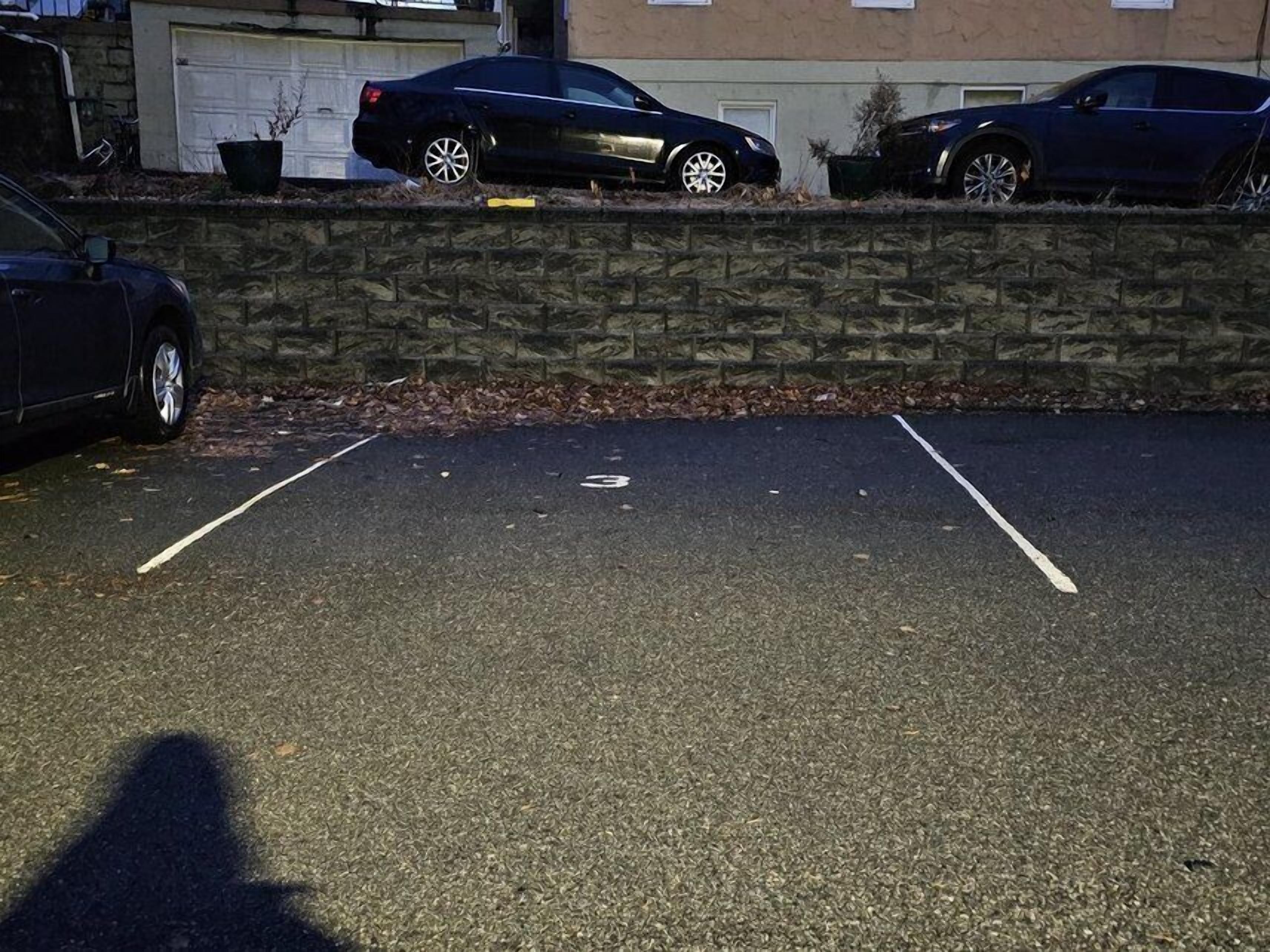 Un parking avec deux voitures garées, un mur de pierre et un bâtiment avec un garage en arrière-plan.
