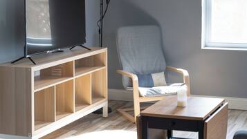Ein modernes Wohnzimmer mit Fernseher auf einem Holzständer, einem grauen Sessel, einem kleinen Holztisch und einem Fenster, das für natürliches Licht sorgt.