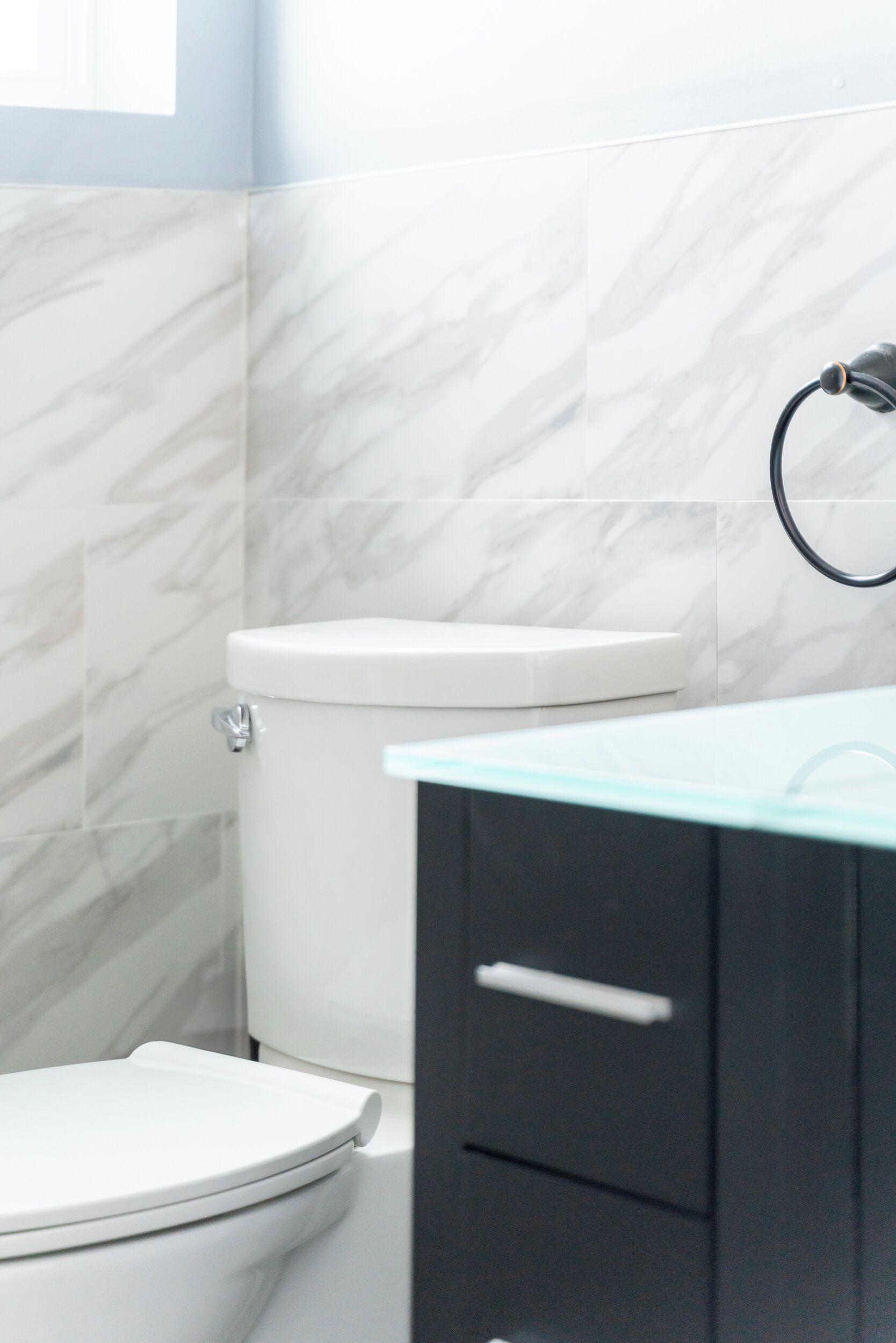 A bathroom with a white toilet, marble tiles, a black cabinet, and a glass countertop.