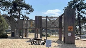 A wooden structure with benches and picnic tables in a sandy area, surrounded by trees.