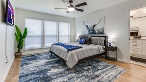 A bedroom with a bed, a ceiling fan, a large window with blinds, a wall-mounted TV, a plant, and a painting.