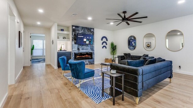 A modern living room with a blue sofa, a blue armchair, and a small table. There is a ceiling fan, a TV, and a kitchen area in the background.