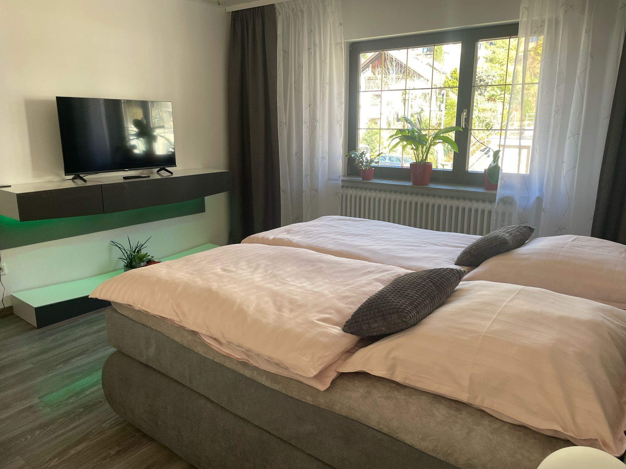 A bedroom with two beds, a TV on a built-in shelf, a window with curtains, and a radiator.