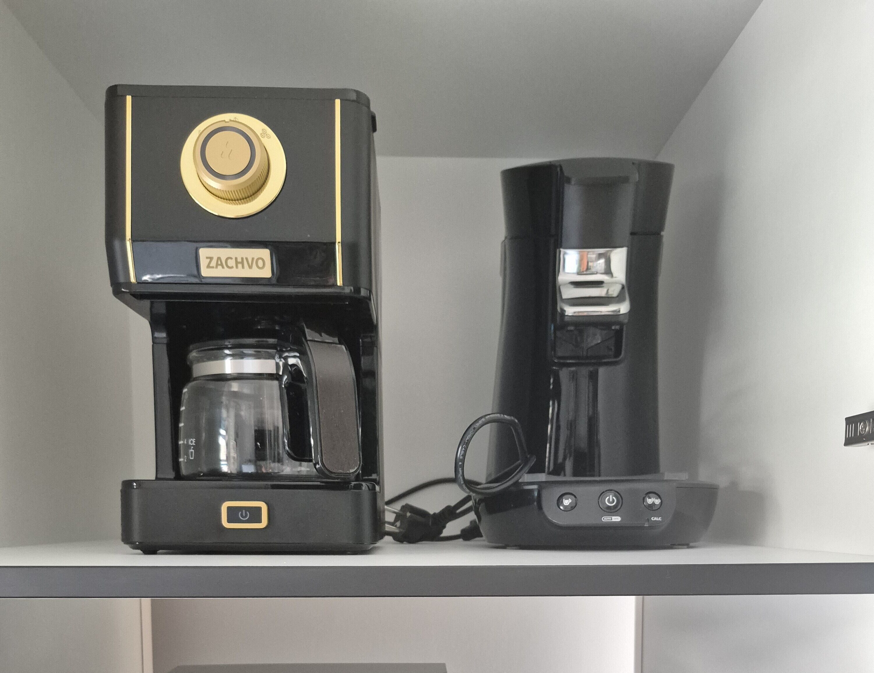 Una cafetera negra con un dial dorado y la etiqueta que dice "ZACHVO" sobre un estante blanco.