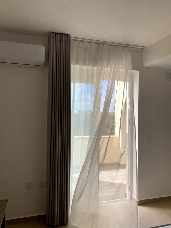 A room with a large sliding glass door leading to a balcony, a tall air conditioning unit on the wall, and a power outlet on the floor.