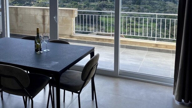 Un balcon spacieux avec une table et des chaises, une bouteille de vin et un verre. Le balcon offre une vue panoramique sur un paysage vallonné, avec des bâtiments et des grues de construction visibles au loin.