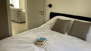 A neatly made bed with white bedding and two pillows, a book on the bed, and a door leading to another room. - The Valley Collection - Suite B23 (Ix-Xagħra)