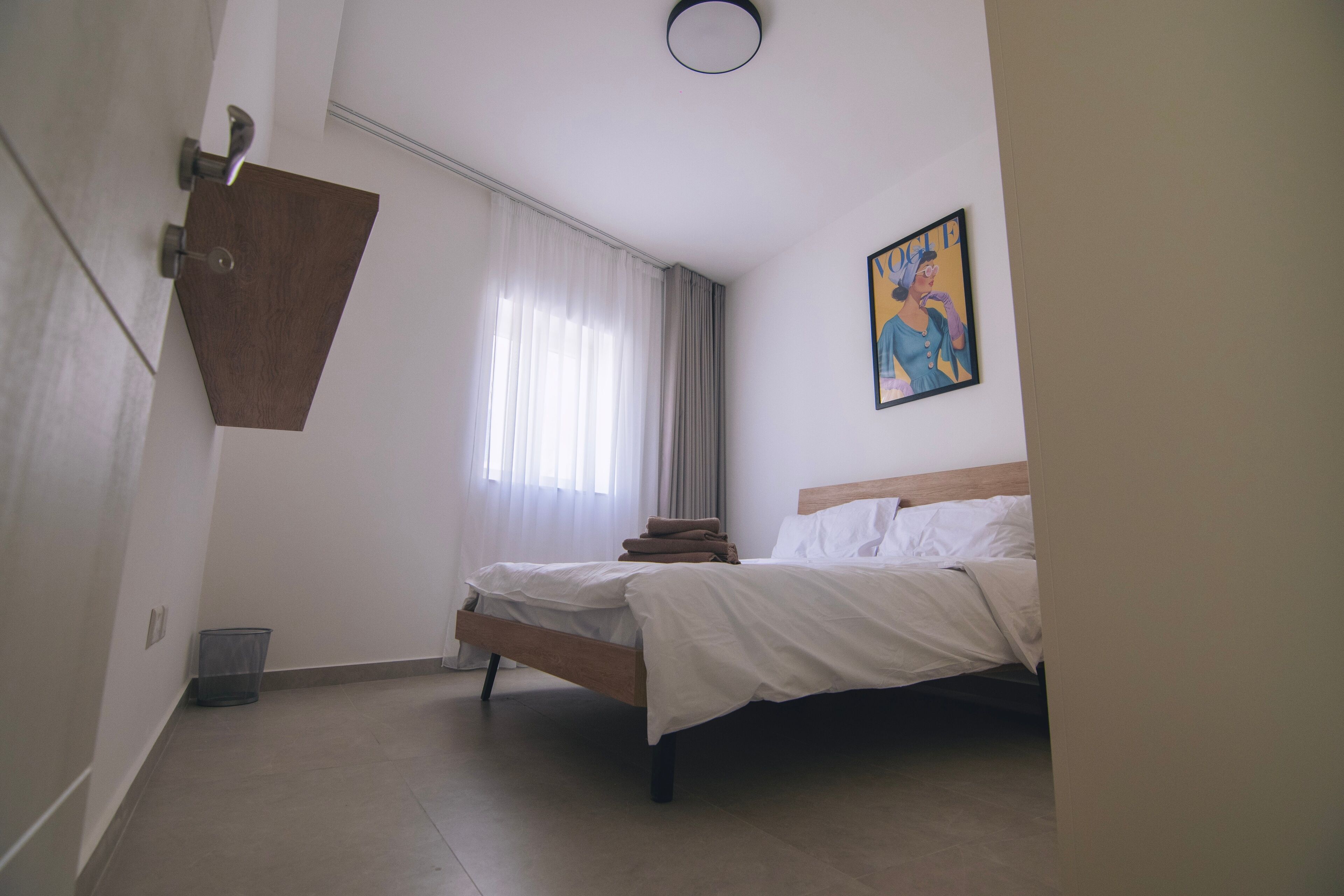 Une chambre à coucher avec un lit, une fenêtre avec des rideaux, un tableau au mur et une poubelle.