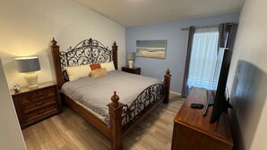 Ein Schlafzimmer mit einem hölzernen Bett, Nachttischen, einer Lampe, einem Fernseher und einem Fenster mit Vorhängen.