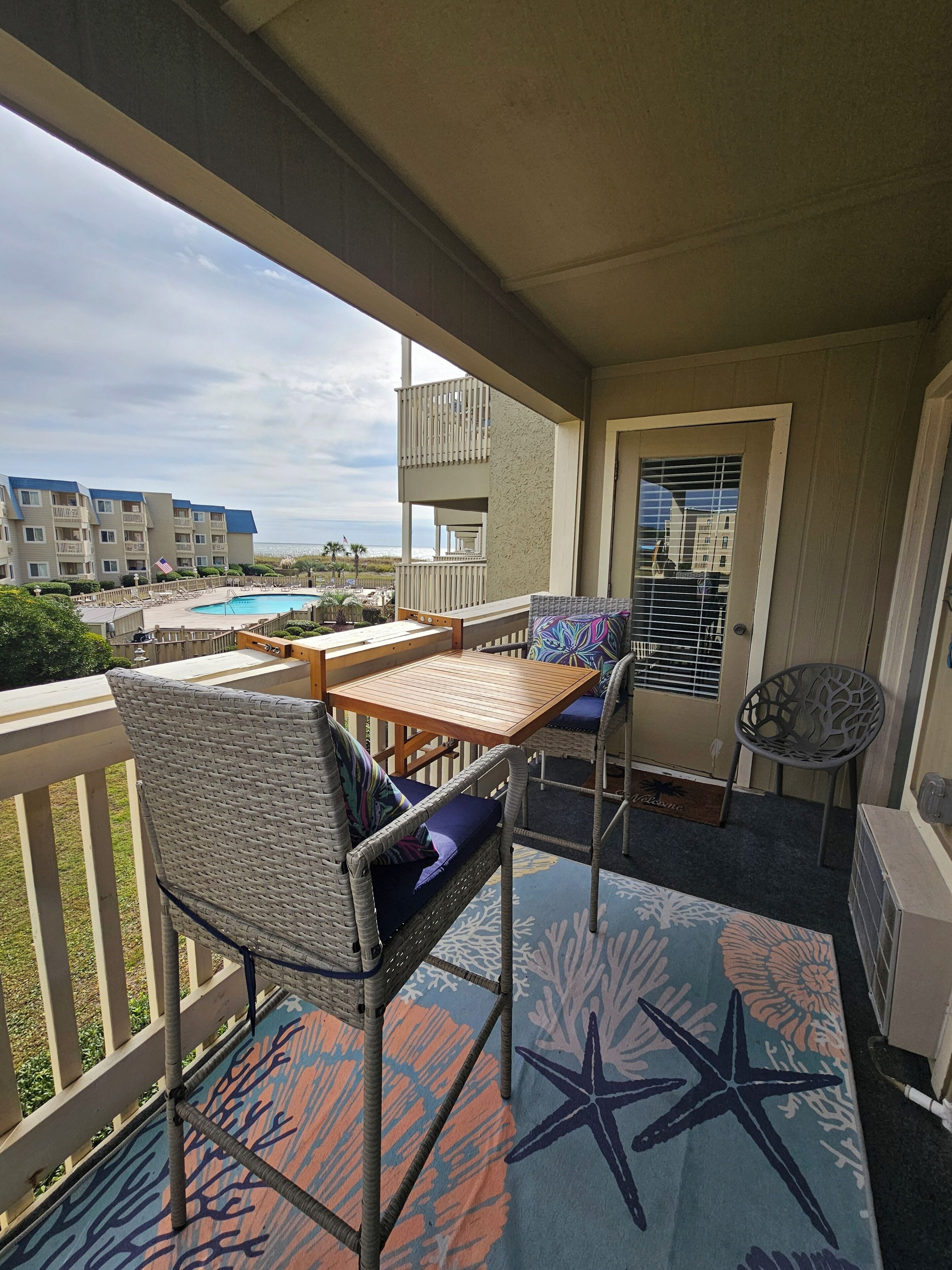 Een balkon met een houten tafel en rieten stoelen, met uitzicht op een zwembad en gebouwen.