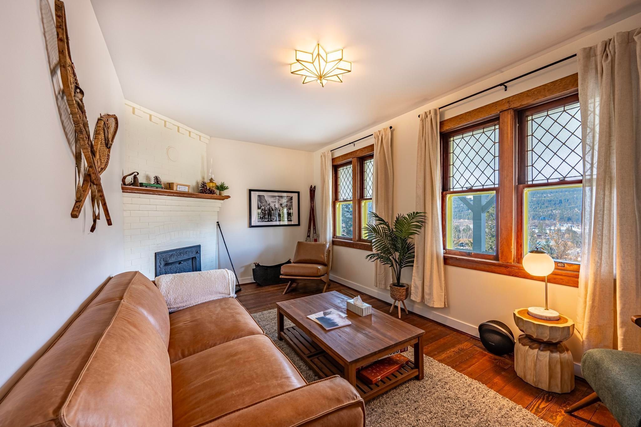 A cozy living room with a brown leather sofa, a wooden coffee table, and a fireplace. There is a large window with curtains and a view of the city.