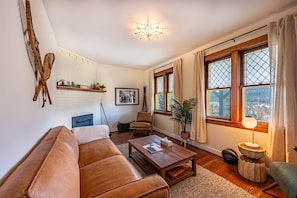 Een gezellige woonkamer met een bruine leren bank, een houten salontafel en een open haard. Er is een groot raam met gordijnen en uitzicht op de stad.