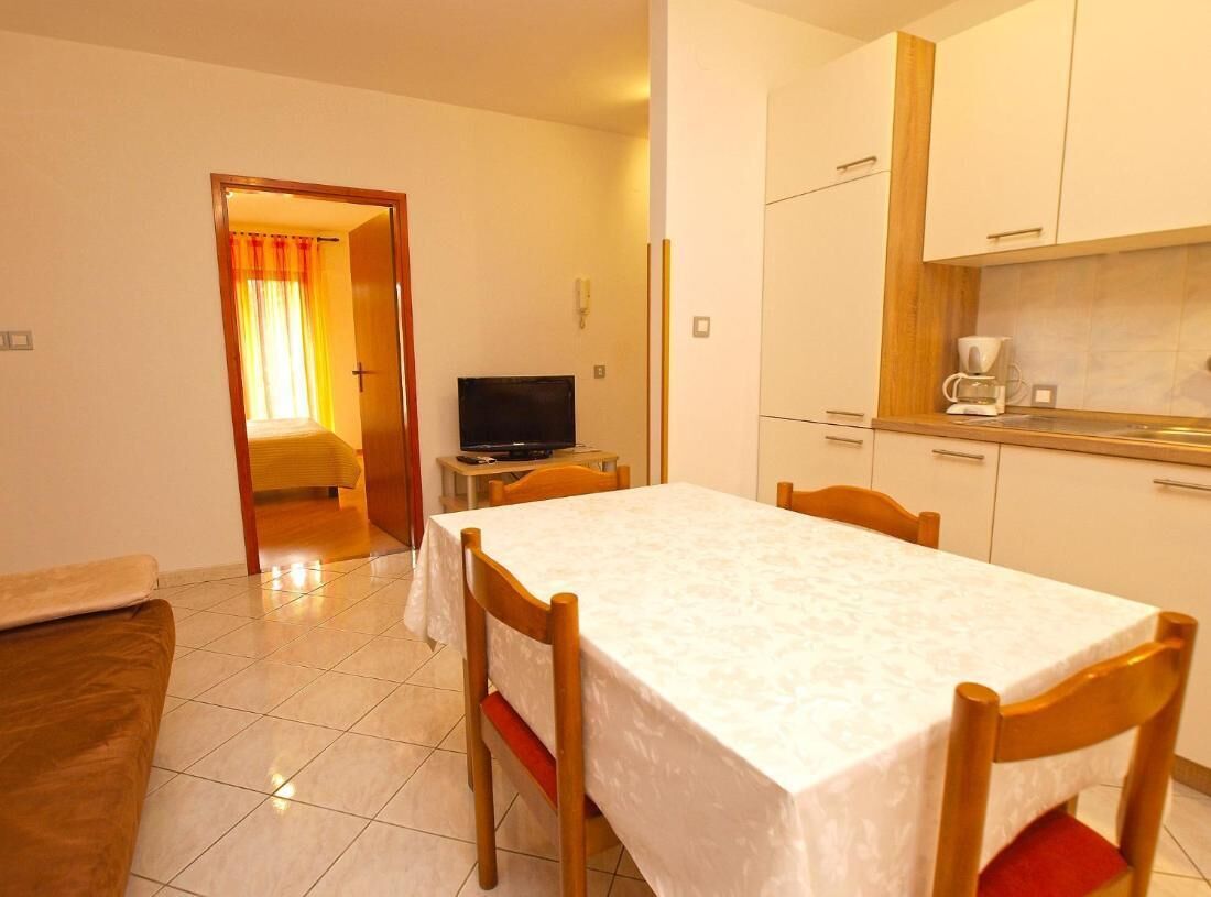 A small, well-lit room with a dining table, chairs, a television, and a kitchenette.