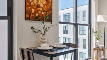 Uma sala de jantar moderna com uma mesa e cadeiras, uma janela ampla com vista para edifícios e uma peça decorativa na parede.