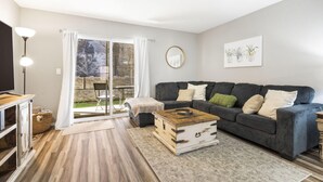 Una sala de estar con un sofá gris oscuro, una mesa de centro de madera y un ventanal con cortinas blancas.