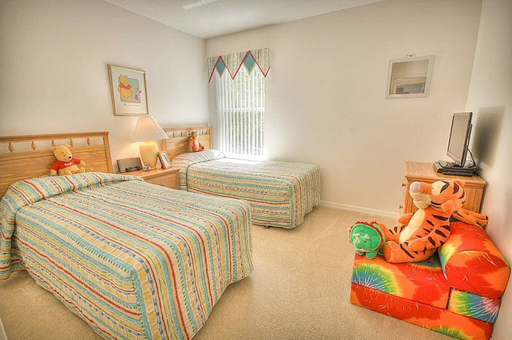 A bedroom with two beds, a nightstand with a lamp, a window with curtains, a framed picture on the wall, and a stuffed toy on the floor.