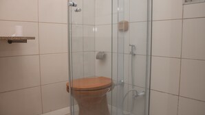 A bathroom with a glass-enclosed shower, a wooden toilet, and a shelf with a roll of toilet paper. - Comfort Suite 203 (Centro Diamantina)