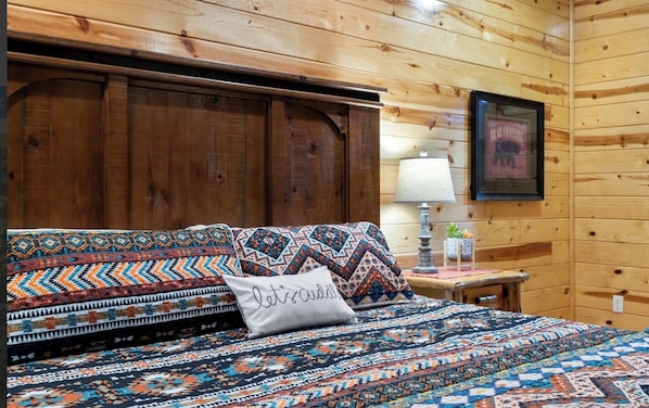Ein Schlafzimmer einer Holzhütte mit einem Bett, Kissen, einem Nachttisch, einer Lampe und einem gerahmten Bild an der Wand.