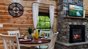 A wooden cabin interior with a dining table set for breakfast, a stone fireplace, and a clock on the wall.