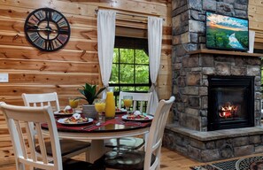 A wooden cabin interior with a dining table set for breakfast, a stone fireplace, and a clock on the wall.