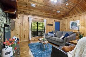 A cozy cabin interior with a stone fireplace, wooden walls, a grey sofa, and a blue rug.