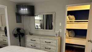 Un dormitorio con cama, cómoda, espejo, televisor, ventilador y una cama litera.