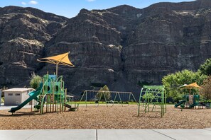 A playground with climbing structures and slides, set against a backdrop of rocky cliffs.