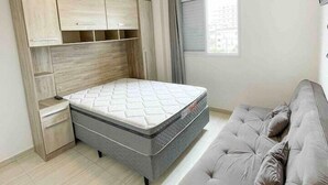 A bedroom with a bed, a grey sofa, a ceiling fan, a window, and a TV on the wall. - Pé na areia apto c vista mar e piscina (Praia Grande)