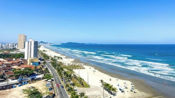 A coastal cityscape with a beach, high-rise buildings, and a clear blue sky. - Pé na areia apto c vista mar e piscina (Praia Grande)