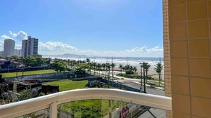 A balcony with a view of a beach, palm trees, and buildings in the distance. - Pé na areia apto c vista mar e piscina (Praia Grande)