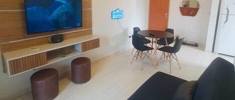 Una sala moderna con televisor de pantalla plana, un mueble bajo para la televisión de madera, sofá negro, mesa de comedor con sillas y una pequeña área de cocina con refrigerador.