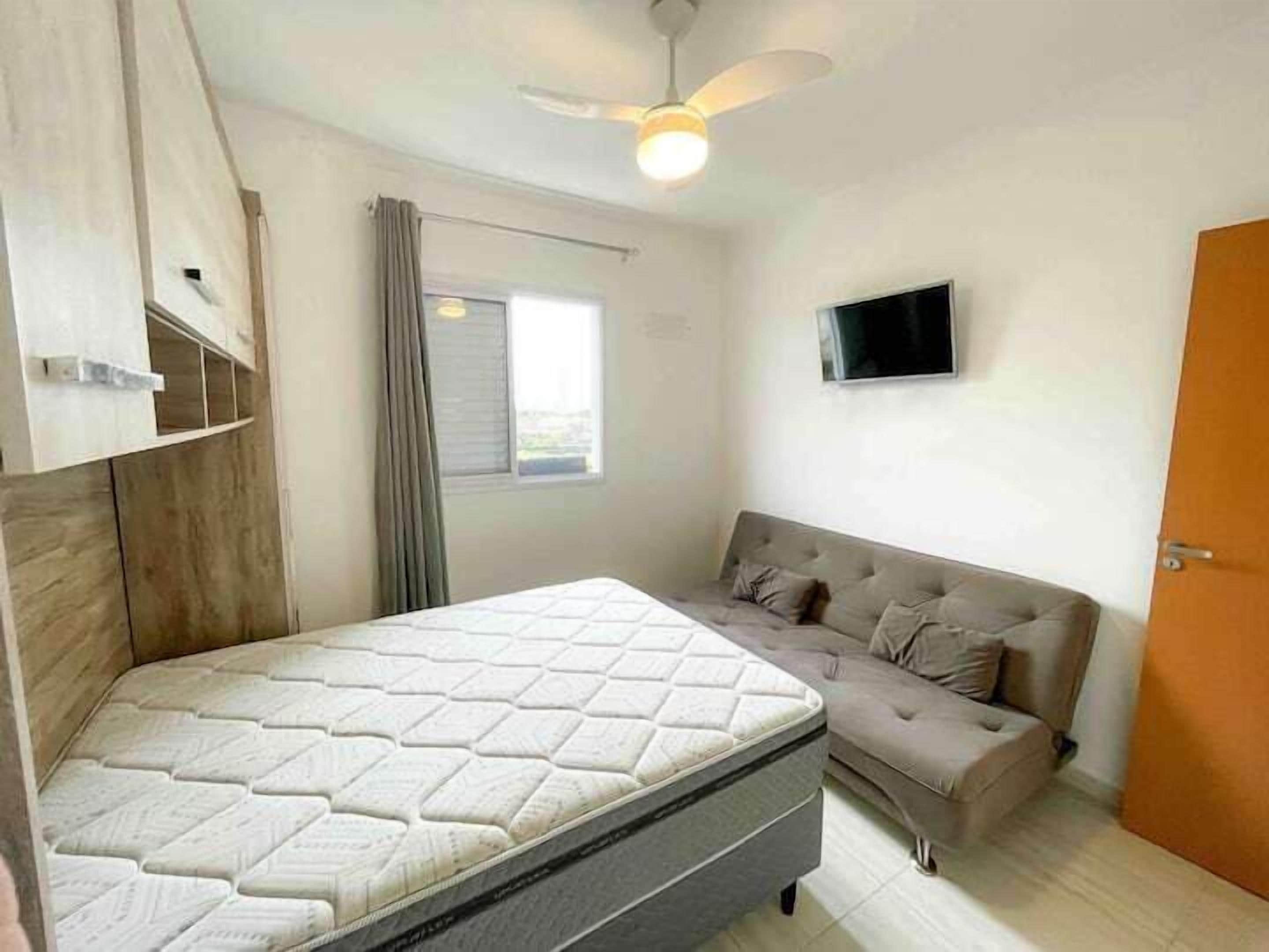 A bedroom with a bed, a grey sofa, a television on the wall, and a window with curtains.