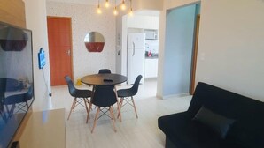A modern living room with a black sofa, a dining table with chairs, a TV, and a kitchen area in the background. - Pé na areia apto c vista mar e piscina (Praia Grande)